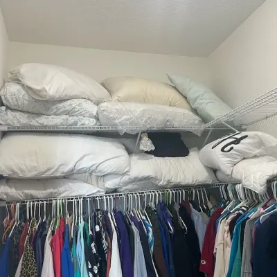 Closet filled with various bed pillows on upper wire shelving and a rack of assorted casual clothing hanging below.