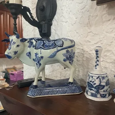 Delft-style blue and white ceramic cow figurine displayed alongside a matching floral porcelain bell on a wooden mantle.