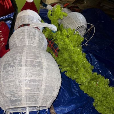 Sale Photo Thumbnail #43: Two collapsible light-up snowman outdoor decorations with fabric bodies, hats, and scarves, shown alongside a green artificial pine garland on a blue tarp.