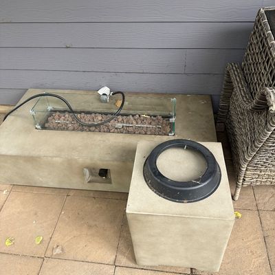 Sale Photo Thumbnail #53: Modern concrete-style rectangular gas fire pit table with a glass wind guard and lava rocks, accompanied by a matching square concrete-style propane tank cover.