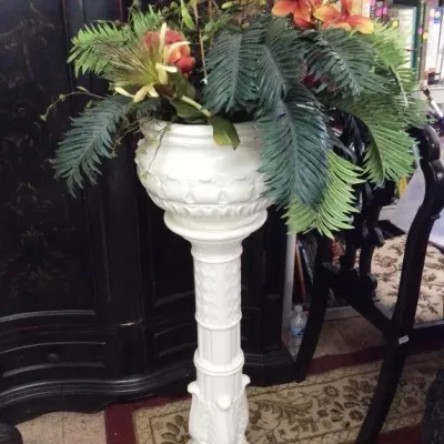 White ceramic pedestal planter featuring a classical column design, filled with an assortment of faux tropical greenery and orange flowers.
