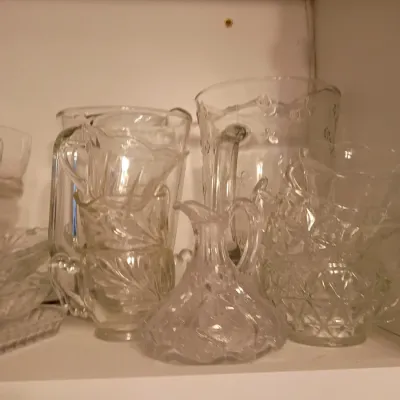 Sale Photo Thumbnail #67: Collection of vintage clear pressed glassware, including water pitchers, creamers, sugar bowls, and a small decanter displayed on a cabinet shelf.