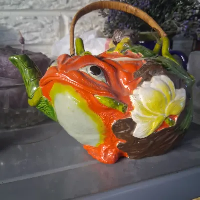 Sale Photo Thumbnail #14: Whimsical ceramic frog-shaped teapot with vibrant orange and green glazes, a molded water lily accent, and a wicker handle.