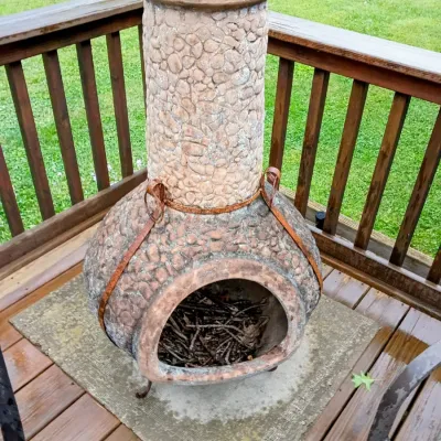 Sale Photo Thumbnail #33: Rustic clay chiminea with pebble-textured finish and a rusted iron stand, displayed on a wooden deck.