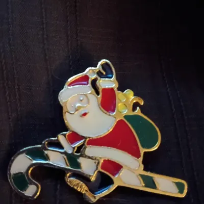 Festive Santa Claus enamel lapel pin featuring Santa riding a candy cane with a toy sack, set in gold-tone metal.