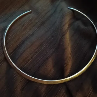 Simple silver-tone metal collar necklace featuring a smooth, open-cuff design resting on a dark textured fabric.