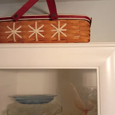 Sale Photo Thumbnail #36: Longaberger woven wood basket featuring cream starburst or snowflake accents, red handles, and trim, perched atop a white cabinet displaying glass tableware.
