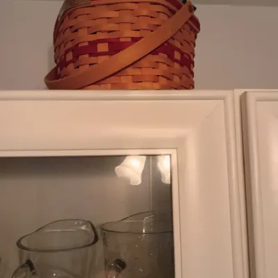 Sale Photo Thumbnail #45: Longaberger woven basket with red accents and a leather handle, displayed on top of a white glass-front cabinet housing clear glass pitchers.