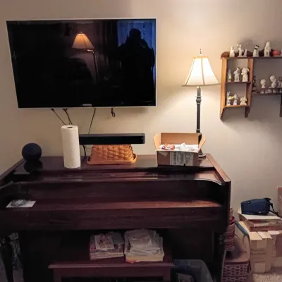 Sale Photo Thumbnail #53: Dark wood spinet piano paired with a wall-mounted flat-screen television and a wooden display shelf containing figurine collections.