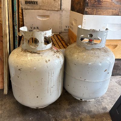Sale Photo Thumbnail #56: Two standard 20-pound propane tanks showing some dirt and wear, positioned in front of stacked wooden beehive boxes.