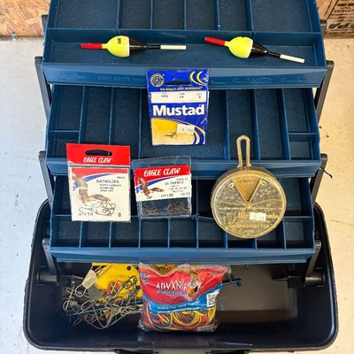 Sale Photo Thumbnail #46: Open blue plastic cantilever tackle box filled with assorted fishing supplies, including Eagle Claw and Mustad hook packs, bobbers, a sinker selector disc, and a fish stringer.