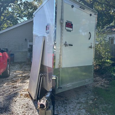 Sale Photo Thumbnail #70: White enclosed cargo trailer featuring chrome trim, diamond plate base accents, rear swing door, and front tongue jack, shown parked on a gravel driveway.