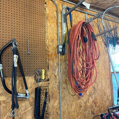 Sale Photo Thumbnail #6: Workshop wall featuring a coiled orange extension cord hanging on a rack, alongside a grease gun, bow saw frame, and various tools.