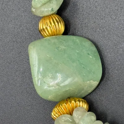 Sale Photo Thumbnail #345: Close-up of a gemstone necklace featuring light green aventurine tumbled chips and a central focal stone flanked by ribbed gold-tone spacer beads.