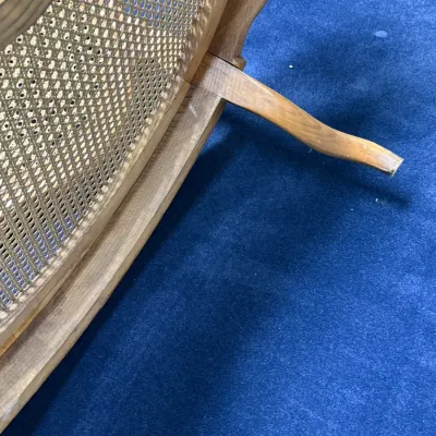 Sale Photo Thumbnail #679: Detail of a vintage cane-back chair showing the woven mesh panel and carved wooden leg resting on blue carpeting.