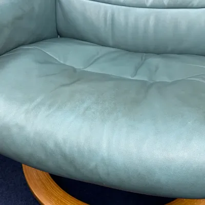 Sale Photo Thumbnail #228: Ekornes Stressless reclining armchair upholstered in light teal leather, featuring a wood ring base and showing some surface wear and discoloration on the seat and armrest.