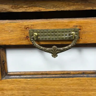 Sale Photo Thumbnail #280: Detail view of an antique wooden drawer featuring a white label window frame and an ornate brass bail pull with a checkered backplate.