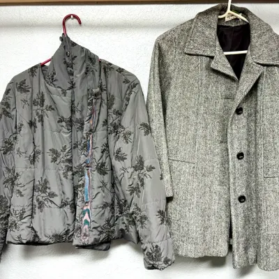 Sale Photo Thumbnail #96: Pair of women's coats hanging on plastic hangers, including a grey floral embroidered quilted jacket and a classic grey herringbone wool outerwear coat.