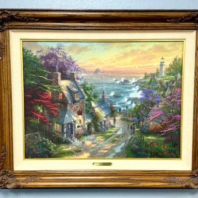 Sale Photo Thumbnail #821: Framed canvas print by Thomas Kinkade titled 'The Village Lighthouse', featuring a coastal village scene housed in an ornate wooden frame with a fabric liner.