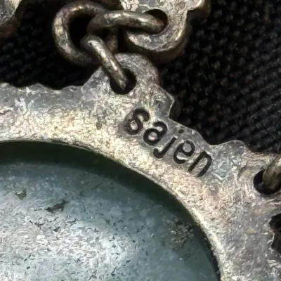 Sale Photo Thumbnail #330: Extreme close-up of a Sajen sterling silver jewelry piece, showing the maker's mark stamped into the metal casing around a green cabochon.