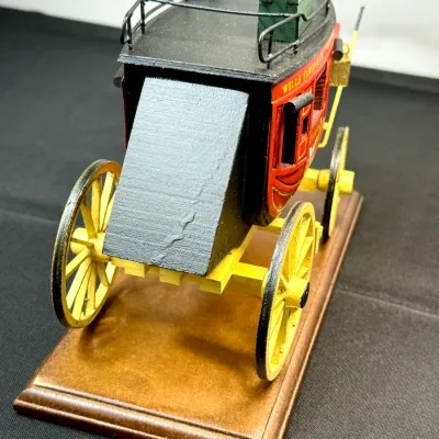 Sale Photo Thumbnail #491: Handcrafted miniature Wells Fargo & Co. stagecoach model, mounted on a wooden display base with painted yellow wheels and red body detailing.