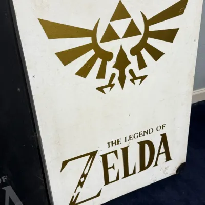 Sale Photo Thumbnail #922: Hand-painted or printed wood panel sign featuring The Legend of Zelda Royal Crest and logo in gold against a white background.