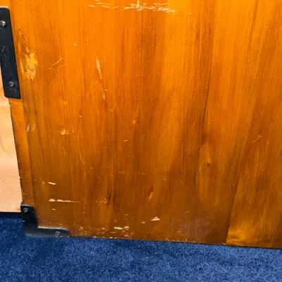 Sale Photo Thumbnail #214: Close-up of a warm-toned wooden cabinet or door showing surface scratches, scuffs, and black metal corner brackets.