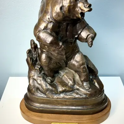 Sale Photo Thumbnail #125: Bronze sculpture of a snarling grizzly bear on a rocky terrain base by James L. Clark, mounted on an oval wooden plinth with a named brass plaque.