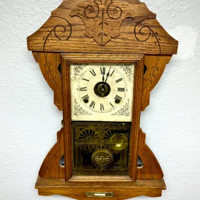Sale Photo Thumbnail #365: Antique Seth Thomas gingerbread wall clock in an oak case featuring carved scroll details and an ornate brass pendulum.