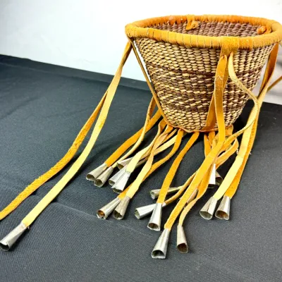 Sale Photo Thumbnail #406: Handwoven Native American-style basket featuring tan buckskin fringe adorned with metal tin cones along the rim.