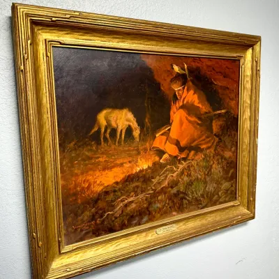 Sale Photo Thumbnail #101: Framed Western oil painting depicting a Native American figure seated by a campfire with a grazing horse in the background. Housed in an ornate gilded wood frame with a title plaque attached to the bottom molding.