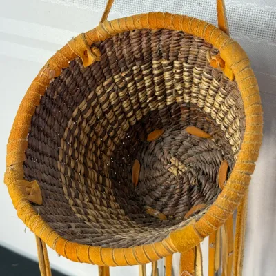 Sale Photo Thumbnail #405: Handwoven Native American-style conical basket featuring a wrapped buckskin rim, leather hanging strap, and leather fringe tassels with metal cones.