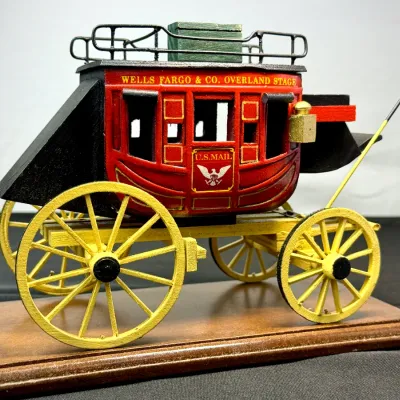 Sale Photo Thumbnail #492: Vintage painted wooden model of a Wells Fargo & Co. Overland Stage stagecoach mounted on a display base, featuring intricate detailing and yellow spoke wheels.