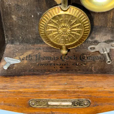 Sale Photo Thumbnail #371: Detail view of a Seth Thomas Clock Company wooden clock interior, featuring a brass pendulum bob, chime bell, original manufacturer's label, and two winding keys.