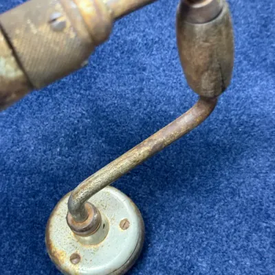 Sale Photo Thumbnail #567: Vintage hand brace drill featuring a worn wooden sweep handle and a circular head unit. The metal frame shows significant rust, patina, and age-related wear.