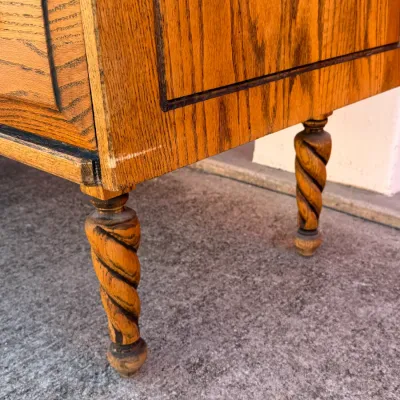 Sale Photo Thumbnail #262: Close-up of an oak wooden cabinet or dresser featuring barley twist legs and panel details, displaying light surface wear along the edges.