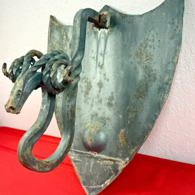 Sale Photo Thumbnail #413: Vintage wrought iron ram's head wall-mounted sconce or bracket, featuring twisted iron horns and an elongated metal shield backing with weathered patina.