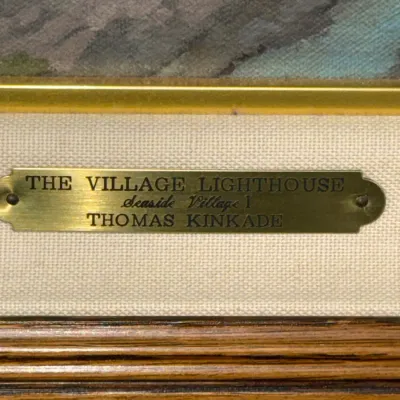 Sale Photo Thumbnail #826: Detail view of a framed Thomas Kinkade artwork titled 'The Village Lighthouse, Seaside Village I', showing the brass title plaque on the matting.