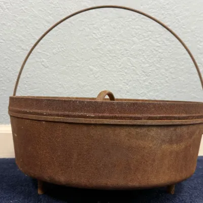 Sale Photo Thumbnail #588: Vintage three-legged cast iron Dutch oven with lid and wire bail handle, showing a heavy rusted patina throughout.