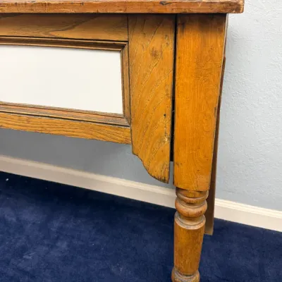 Sale Photo Thumbnail #288: Detail view of an antique wooden table or desk featuring turned legs, a framed front inset panel, and a rustic distressed finish.