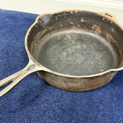 Sale Photo Thumbnail #583: Vintage cast iron skillet with dual pour spouts and a helper handle, showing surface wear and minor rust consistent with age.