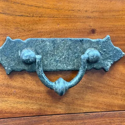 Sale Photo Thumbnail #248: Rustic weathered metal drawer pull with an ornate backplate, mounted on a polished wood cabinet drawer.