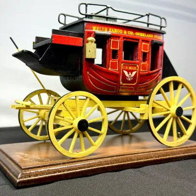 Sale Photo Thumbnail #490: Miniature wooden Wells Fargo & Co. Overland Stagecoach model mounted on a display base, featuring painted red, yellow, and black details.