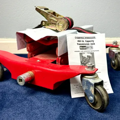 Sale Photo Thumbnail #525: Central Hydraulics 450 lb. capacity transmission jack, Model 39178, featuring a red low-profile frame with caster wheels, ratchet strap, and original instruction manual.