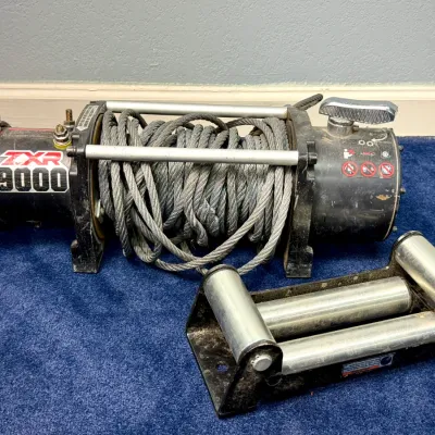 Sale Photo Thumbnail #60: Badland ZXR 9000-lb capacity electric truck winch with steel cable and detached roller fairlead. Shows light surface wear and dirt from prior use.