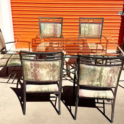 Sale Photo Thumbnail #17: Seven-piece outdoor patio dining set featuring a rectangular glass-top table and six dark metal chairs with patterned cushions.