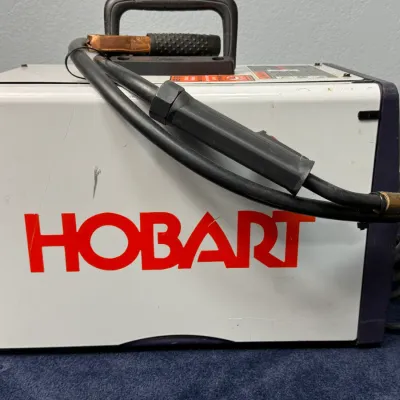 Sale Photo Thumbnail #500: Hobart portable MIG welder with attached gun, ground clamp, and top carrying handle, showing light exterior scuffs from use.