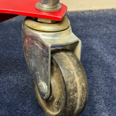 Sale Photo Thumbnail #527: Close-up of a heavy-duty swivel caster wheel attached to a red metal base, showing minor wear and surface dirt.