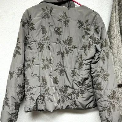 Sale Photo Thumbnail #902: Grey quilted jacket with an embroidered leaf pattern hanging on a hanger.