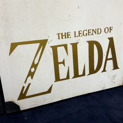 Sale Photo Thumbnail #925: Vintage painted wooden sign featuring 'The Legend of Zelda' logo in gold lettering on a white background with metal corner caps.